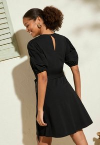 Robe noire avec des manches bouffantes, taille cintrée et jupe évasée. Détail dos en trou de serrure et nœud à la taille. Matière en coton doux.