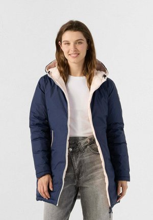 Chaqueta de plumas azul marino con capucha forrada en rosa, cremallera frontal, bolsillos laterales y patrón acolchado. Lleva puesta sobre una camiseta blanca y unos jeans grises.