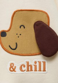 Tessuto beige con un volto di cane di cartone ricamato in marrone e arancione. Sotto il cane è scritto "& chill" in arancione e bianco.
