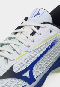 Κοντινή λήψη λευκού αθλητικού παπουτσιού με υφή στη μία πλευρά, μπλε και νεον κίτρινες λεπτομέρειες, λευκά κορδόνια και μαύρη εσωτερική επένδυση.