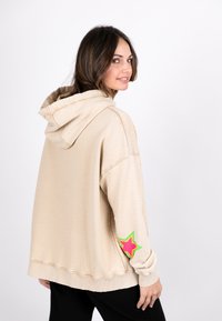 Beige oversized hoodie with a front pocket, drawstring hood, and a colorful star patch on the left sleeve. Soft cotton material.