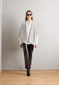 Grå, struktureret poncho med to frontlommer lagdelt over en hvid bluse og skinnende sorte læderbukser, parret med sorte hæle støvler.