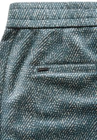 Blauw textiel met een patroon van witte stippen, met een klein rechthoekig label dat aan de achterkant vlakbij een paspelzak is genaaid.