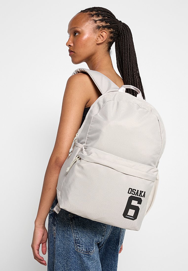 Jonge vrouw met lange vlechten draagt een lichtbeige rugzak met een voorzak met rits en de tekst "OSAKA 6", gecombineerd met blauwe denim jeans.