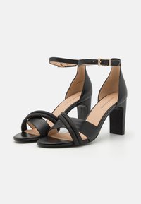 Schwarze High Heels Sandalen mit einem glatten Lederobermaterial, einem umwickelten Knöchelriemen mit einer Schnalle in Goldfarbe und einem strukturierten, überkreuzten Frontband.
