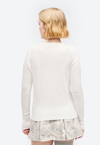 Γυναίκα με κοντά ξανθά μαλλιά, φορώντας ένα λευκό μακρυμάνικο κορμάκι και μια ανοιχτογκρι πλισέ φούστα με λουλουδάτο σχέδιο, γυρισμένη πλάτη.