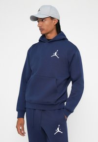 Hanorac bleumarin și pantaloni de trening dintr-un amestec de bumbac. Prezintă o buzunar frontal tip kangur și un logo Jumpman alb pe piept și pe picior. Șapca este de culoare gri deschis.
