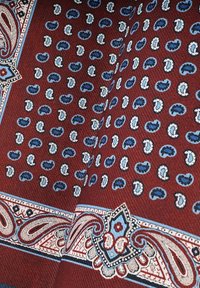 Jedwabna chusta z bordo tłem, ozdobiona niebieskimi i białymi wzorami paisley, otoczona ozdobnymi detalami w kolorze niebieskim i kremowym.