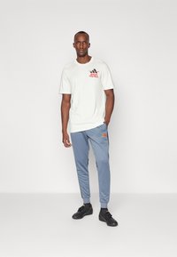 Hvid bomulds-t-shirt med "ADIDAS FOOTBALL" grafik i rød, parret med lyseblå træningsbukser med sorte accenter og et Adidas-logo. Sorte sko.