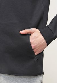 Hand med en liten tatuering delvis inuti dragkedjefickan på en svart sweatshirt som bärs över grå byxor.