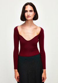Top bordeaux a maniche lunghe con scollo a V profondo, texture a coste, design aderente, abbinato a una gonna scura a righe sottili.