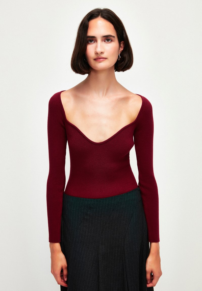 Top bordeaux a maniche lunghe con scollo a V profondo, texture a coste, design aderente, abbinato a una gonna scura a righe sottili.