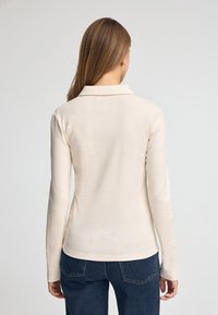 Camicia a maniche lunghe e colletto di colore crema chiaro, caratterizzata da una texture liscia e un design aderente, abbinata a jeans blu scuro.