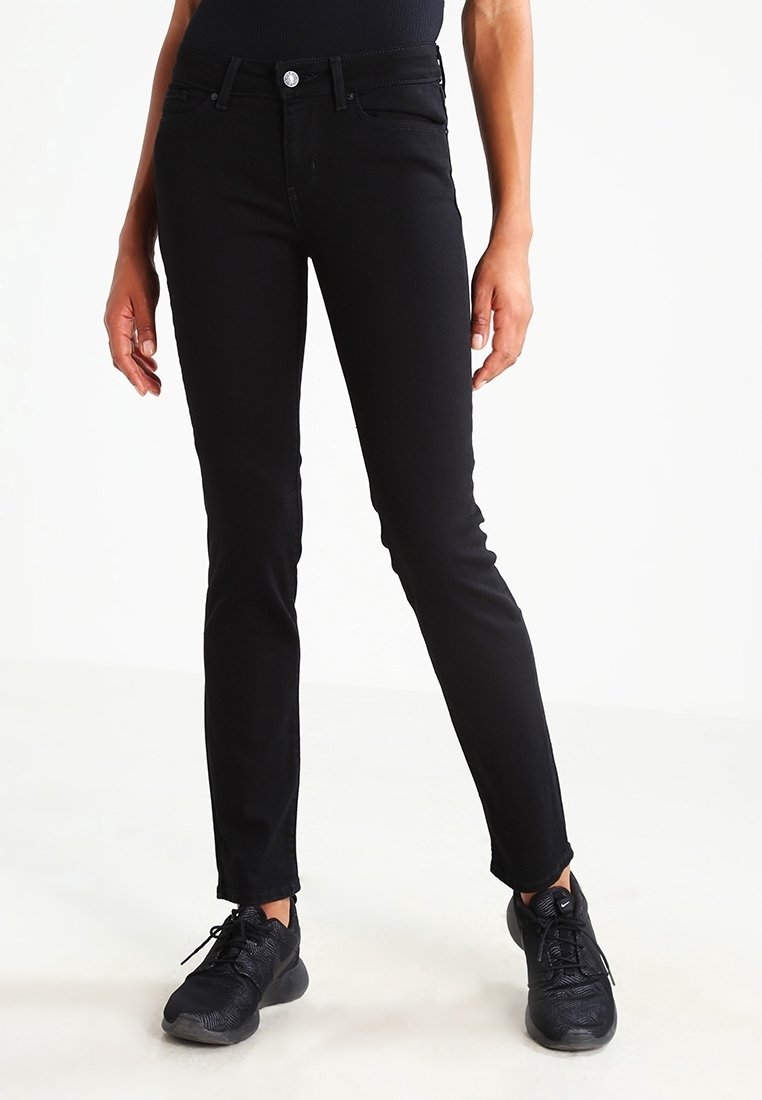 Jean noir slim porté par une personne debout, assorti à des baskets noires, sur fond blanc.