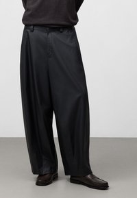 Svarta vida byxor i ett slätt material, med en framfåll, bältesslingor och sidofickor, tillsammans med bruna loafers.