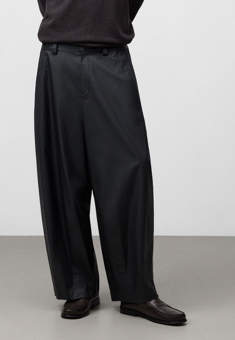 Svarta vida byxor i ett slätt material, med en framfåll, bältesslingor och sidofickor, tillsammans med bruna loafers.