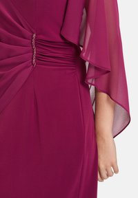 Robe fuchsia avec des manches transparentes fluides, des détails rassemblés sur les côtés et un accent décoratif en cristal à la taille, réalisée dans un tissu lisse et doux.
