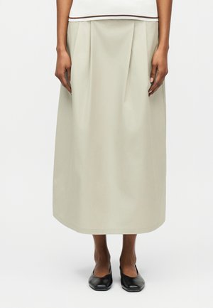 Person iført en beige midi-nederdel med plisseringer, en hvid top med brun kant og sorte flade sko, stående foran en hvid baggrund.