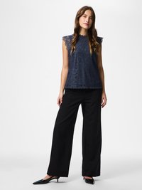 Fiatal nő áll, kezét oldalt tartva, ujjatlan sötétkék csipkefelsőt, fekete bőszárú nadrágot és fekete alacsony sarkú hegyes cipőt visel.