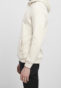 Sweat à capuche beige clair avec cordon de serrage, poche kangourou, poignets et ourlet côtelés. Confectionné dans un mélange de coton doux. Vue de profil.