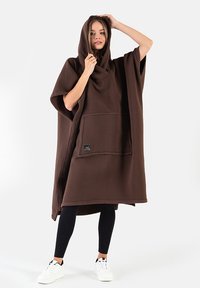 Brązowy polarowy poncho z kapturem, wyposażony w przednią kieszeń i szerokie rękawy. Nosi się go na czarne legginsy i białe sneakersy.