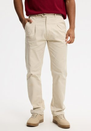 Hombre con pantalones plisados beige, zapatillas beige y una camisa roja con una mano en el bolsillo sobre un fondo blanco.