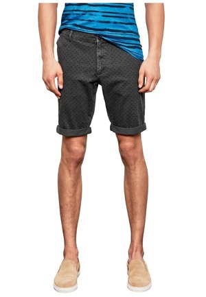 Homme portant un short noir à motifs retroussé, une chemise à rayures bleues et des chaussures beiges sans lacets, debout les mains détendues le long du corps.