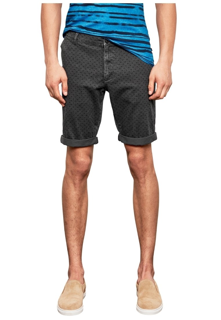 Homme portant un short noir à motifs retroussé, une chemise à rayures bleues et des chaussures beiges sans lacets, debout les mains détendues le long du corps.