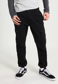 Pantaloni cargo neri con tasche laterali, realizzati in tessuto resistente. Abbinati a sneakers bianche e nere, con bottone frontale e vestibilità rilassata.