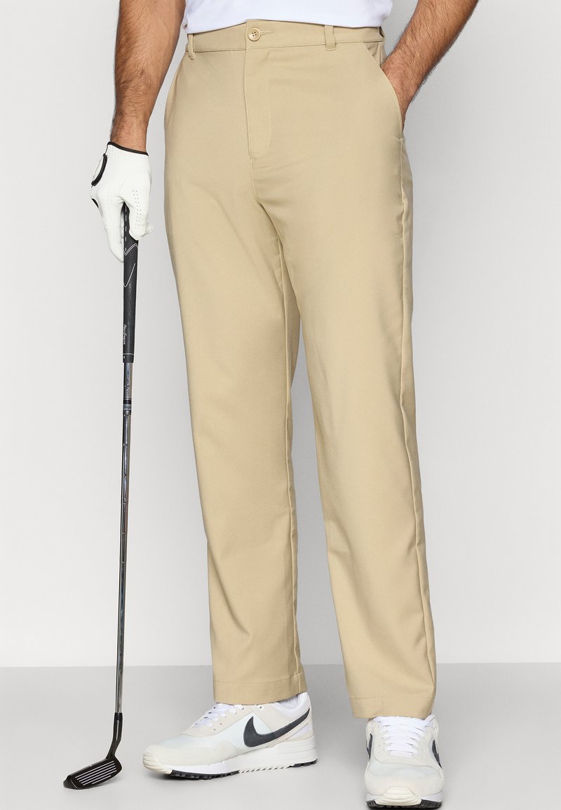 Pantalon de golf beige avec un design à jambe droite, doté d'une fermeture par un seul bouton. Porté avec une chemise blanche et des baskets, tenant un club de golf.