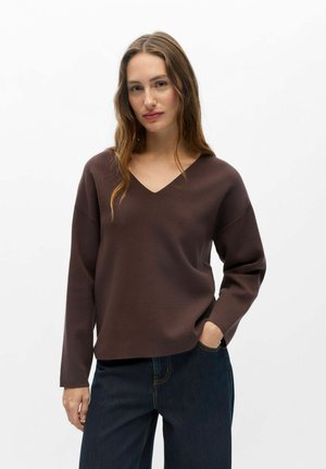 Kvinde med langt bølget hår iført en brun V-hals sweater og mørkeblå jeans, stående med den ene hånd i lommen mod en ensfarvet hvid baggrund.
