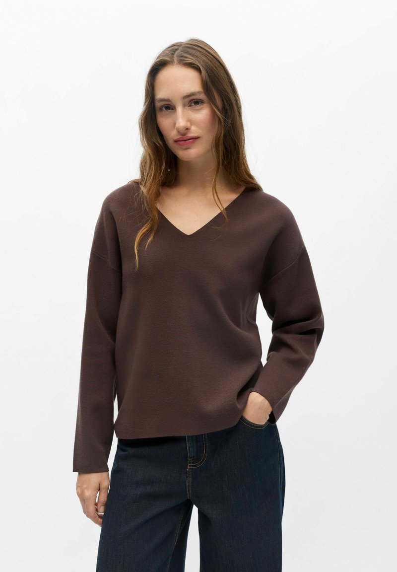Femme aux longs cheveux ondulés porte un pull marron à col en V et un jean bleu foncé, debout avec une main dans la poche devant un fond blanc uni.