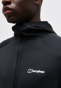 Κοντινό πλάνο ενός ατόμου που φοράει μαύρο μπουφάν Berghaus με φερμουάρ, με το λογότυπο ορατό στο στήθος, σε απλό ανοιχτόχρωμο φόντο.