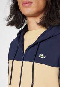 Felpa con zip navy e beige con tasca a marsupio, caratterizzata da un tessuto testurizzato, cappuccio con cordino e un logo verde ricamato sul petto.