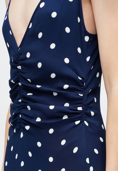 Αμάνικο μπλε φόρεμα με λευκά πουά, βαθύ V λαιμόκοψη και ζάρες ύφασμα κατά μήκος του μπροστινού και των πλευρών.