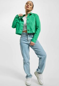 Mujer con chaqueta verde, top corto blanco, vaqueros azul claro y zapatillas blancas posando frente a un fondo blanco liso.