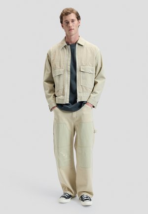 Młody mężczyzna w beżowej kurtce i spodniach z naszywkami, ciemnoszarej koszuli oraz czarno-białych trampkach, stojący z rękami w kieszeniach.