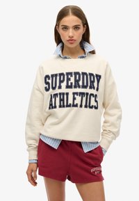 Sudadera crema con estampado en navy "SUPERDRY ATHLETICS", superpuesta a una camisa de cuello a rayas azules, combinada con pantalones cortos color burdeos.