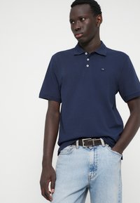 Μπλε βαμβακερό πόλο μπλουζάκι με τρεις κουμπάκια και λεπτομέρεια μικρού λογοτύπου, συνδυασμένο με ανοιχτό τζιν παντελόνι και μαύρη ζώνη.