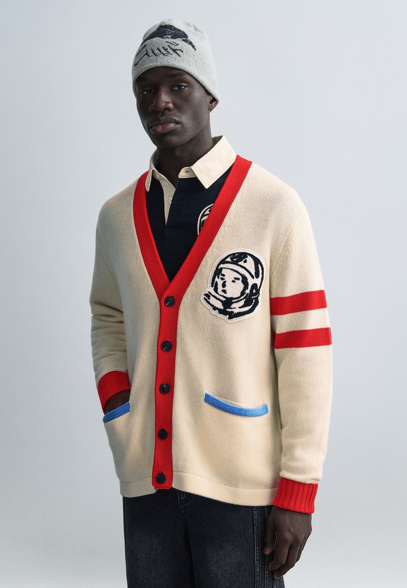 Κρεμ ζακέτα με κόκκινες και μπλε λεπτομέρειες, με μπαλώμα αστροναύτη, μαύρα κουμπιά και αντίθετες πλεκτές μανσέτες. Φορεμένη πάνω από σκούρο πόλο πουκάμισο.