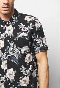 Chemise noire à manches courtes avec un motif floral dans des tons de crème et de vert, dotée d'un col boutonné et d'une poche sur la poitrine.