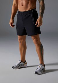 Μαύρο αθλητικό σορτς με ελαστική μέση, πλαϊνή τσέπη με φερμουάρ και εφαρμοστή σχεδίαση. Συνδυάζεται με γκρι και ασημένια παπούτσια για τρέξιμο.