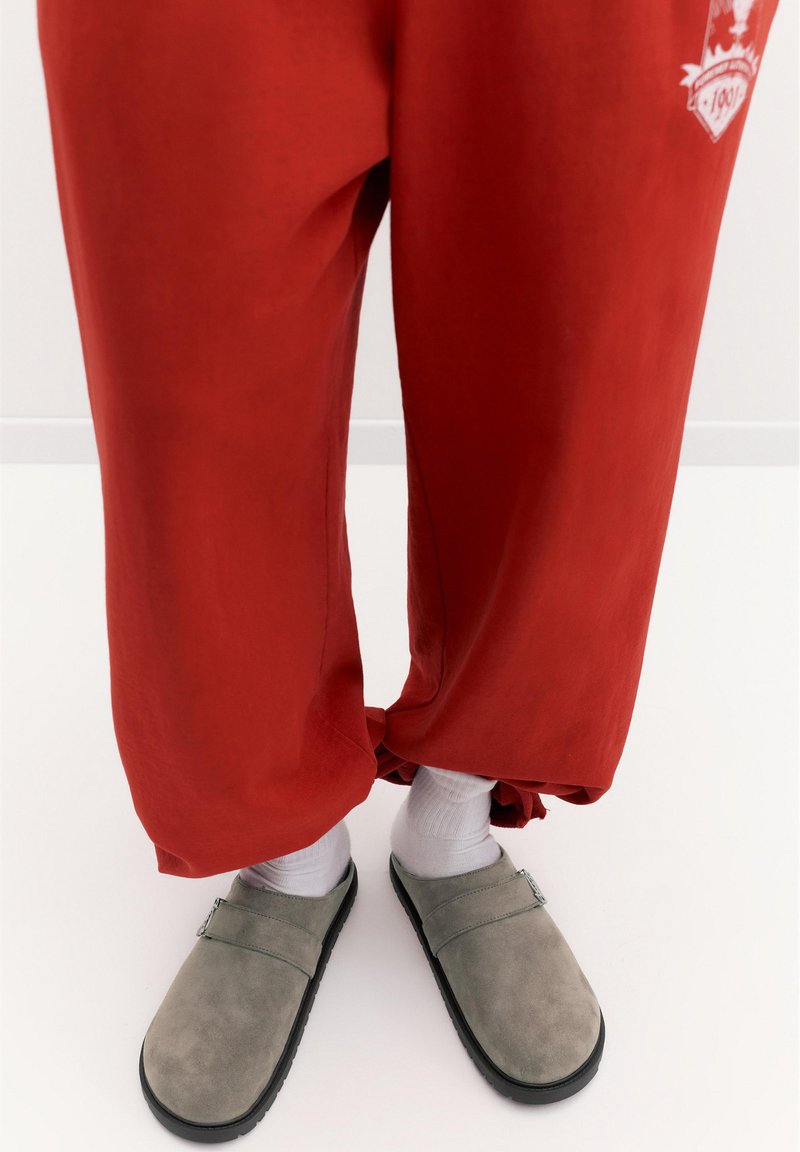 Personne portant un pantalon rouge ample, des chaussettes blanches et des chaussures grises à enfiler avec des semelles noires, debout sur un sol blanc.