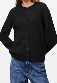 Cárdigan negro de punto con cuello redondo, cierre de botones y mangas largas, combinado con vaqueros de mezclilla azul. Textura suave.