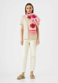 Lichte, roze sjaal met rode rozenpatronen en franjes, gedrapeerd over een beige top en crèmekleurige broek, gecombineerd met geknoopte sandalen.