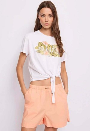 Donna che indossa una t-shirt bianca con nodo frontale, floral e logo, e shorts pesca a vita elastica, in piedi con le mani nelle tasche su sfondo bianco.
