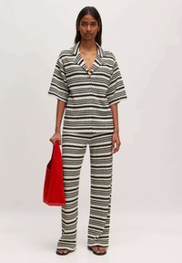 Gestreifter Jumpsuit in Schwarz und Weiß, mit Kragen, kurzen Ärmeln und weitem Bein. Hält eine rote Tragetasche. Flache Sandalen.