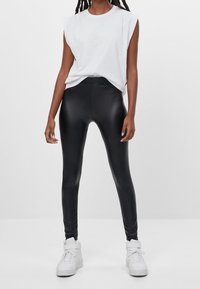 Svarta leggings i konstläder med en slät textur, hög midja, kombinerat med en löst sittande vit ärmlös topp och vita sneakers.