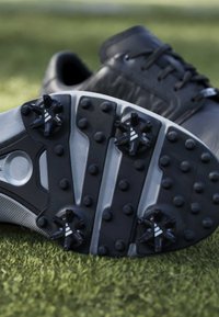 Chaussures de golf noires avec semelles crantées reposant sur de l'herbe verte, mettant en avant les pointes inférieures et le design de la semelle.