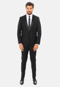 Tuxedo nero con rever in satin, camicia bianca, cravatta nera e pantaloni slim-fit. Scarpe eleganti nere lucide. Sfondo neutro.